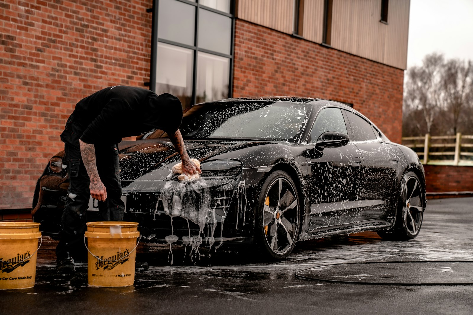A detailer hand-washing a foamed dark sedan with a wash mitt and two buckets.